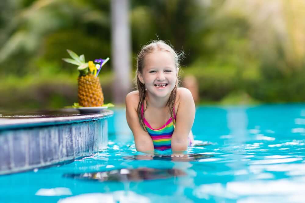 Find out the best Hawaii family resorts with kids club programming by top Hawaii blog Hawaii Travel with Kids. Image of a girl in a swimming pool with a pineapple drink.