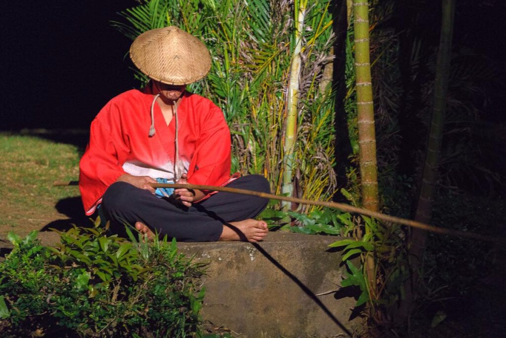 Image of a Japanese man sitting on a rock and fishing with a bamboo pole as part of the Kauai luau entertainment.