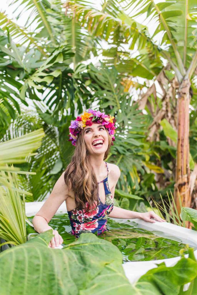 This is one of the most unique Kauai photography sessions you can book. Image of a woman wearing a tropical swimsuit in an outdoor bathtub filled with green monstera leaves.