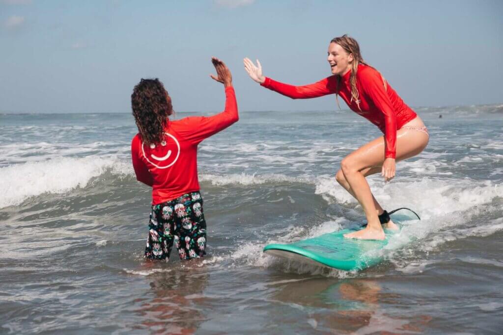 Always wear a rashguard when surfing on Oahu with kids. Image of a woman on a surfboard high fiving her instructor. Both are wearing red long sleeve rash guards.