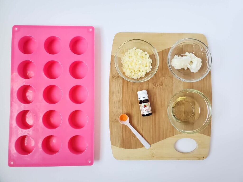 Here's what you need for this Mango Lotion Bar Recipe. Image of a pink silicone mold, fragrance, beeswax, a spoon, on a wood cutting board.