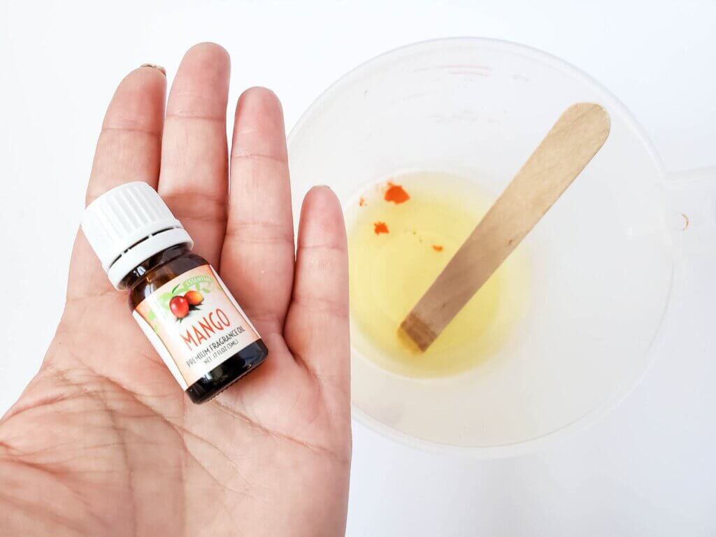 Add in the fragrance and mica powder to create this Hawaiian lotion bar. Image of a woman holding a bottle of mango fragrance in front of a glass bowl.