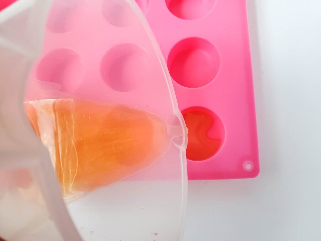 Pour the mango lotion mixture into the silicone molds. Image of someone pouring orange liquid into a pink silicone soap mold.