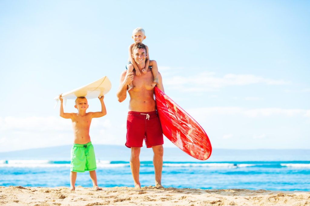 Find out where to book Maui surfing lessons for kids recommended by top Hawaii blog Hawaii Travel with Kids. Image of a father and young sons going surfing at the beach.