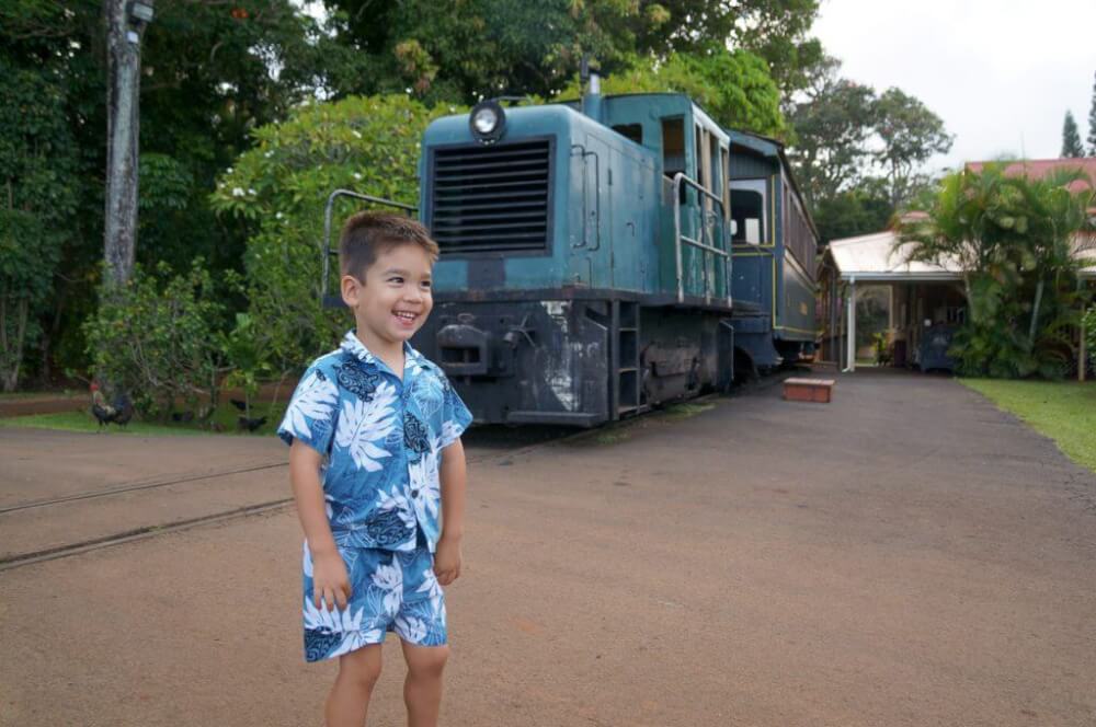 Image of a boy wearing an Aloha shirt and shorts posing in front of a historic train on Kauai.