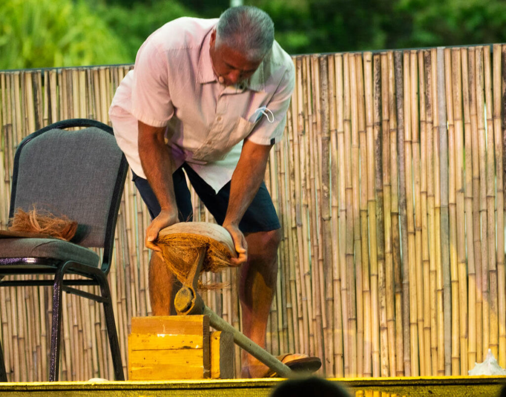 Learn how to husk a coconut at the Ahi Lele Fire Show on Kauai. Image of a man using a tool to break open a coconut on stage.