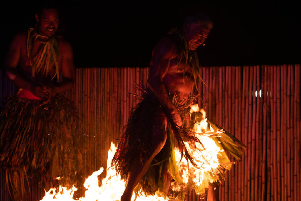 See dancers walk across fire at this Hawaiian fire dancer show. Image of a man wearing a grass skirt walking across fire.