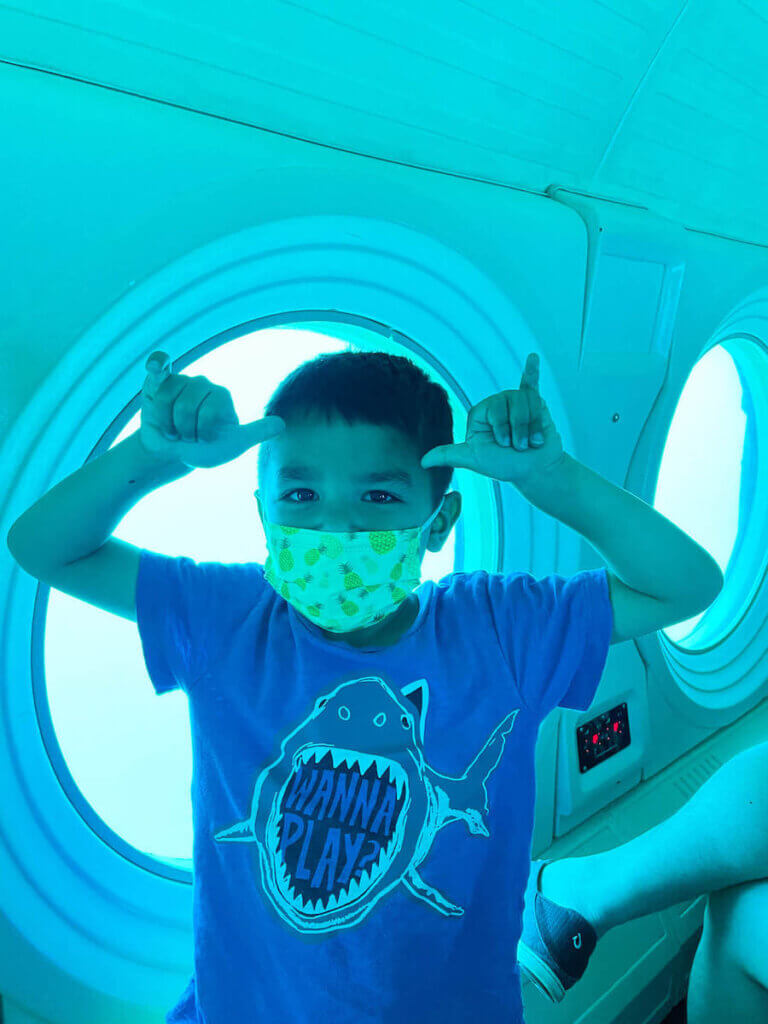 Is this Oahu submarine tour worth it? My kids say YES! Image of a boy wearing a shark shirt throwing 2 shakas in a Waikiki submarine.