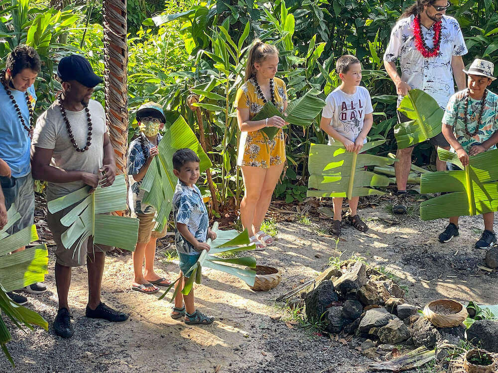 Kids get to participate in activites at Experience Nutridge Luau on Oahu. Image of people holding leaves by an imu on OAhu.