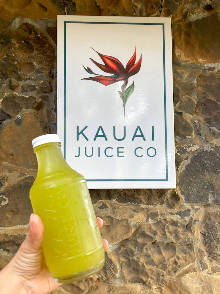 One of the best things to do in Hanalei is drive to Kauai Juice Co in Kilauea for their homebrewed kombucha. Image of a bottle of kombucha in front of the Kauai Juice Co sign.