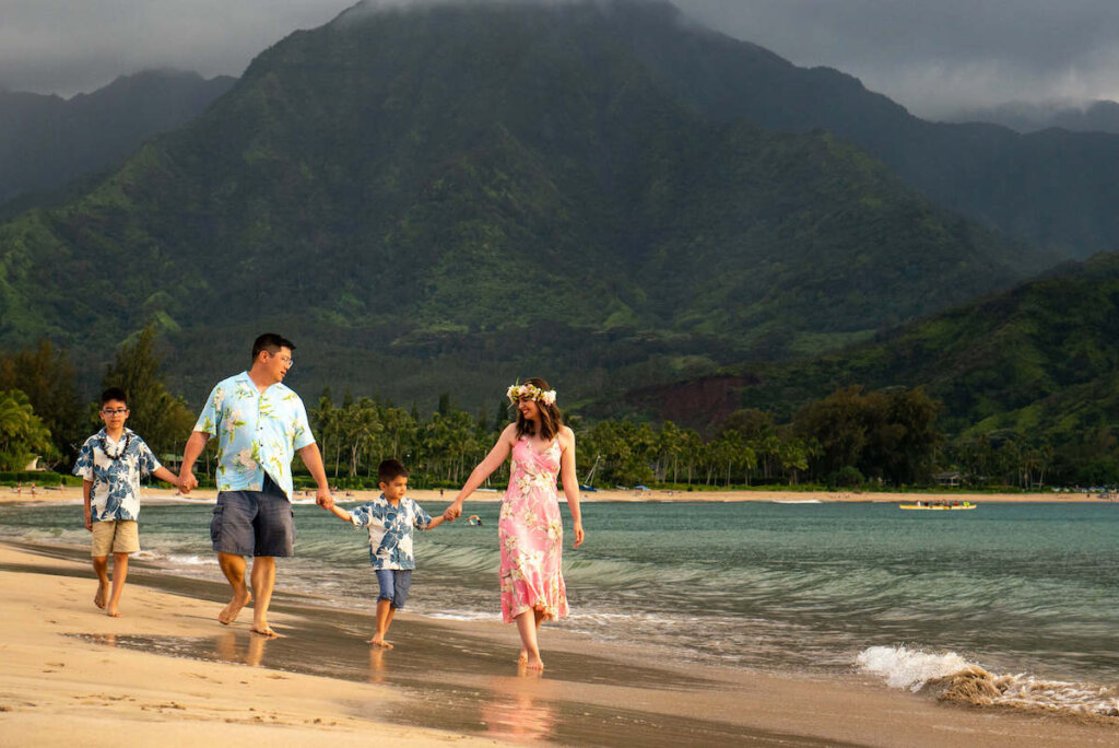 Find out how to book affordable Kauai photographers who will take amazing Hawaii family photos! Image of a family holding hands and walking on the beach on Kauai.