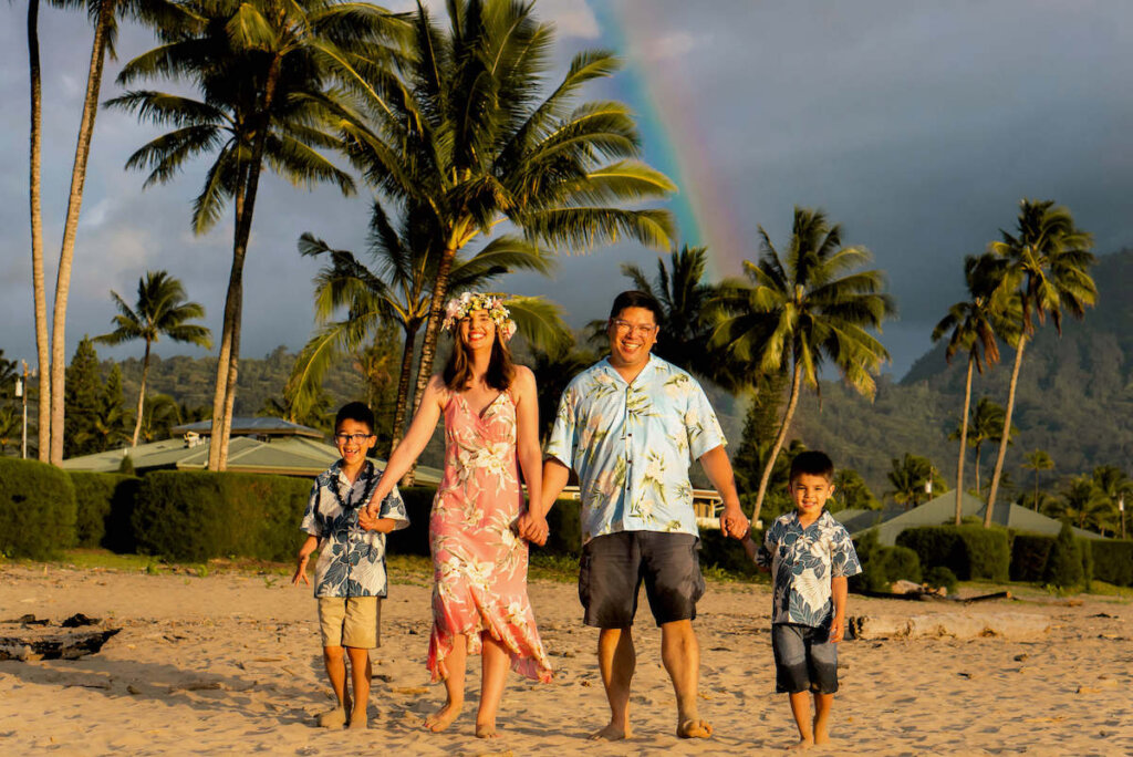 One of my favorite things to do in Hanalei is a professional Kauai photography session. Image of a family wearing Aloha wear smiling and holding hands on Hanalei Bay with a rainbow in the background.