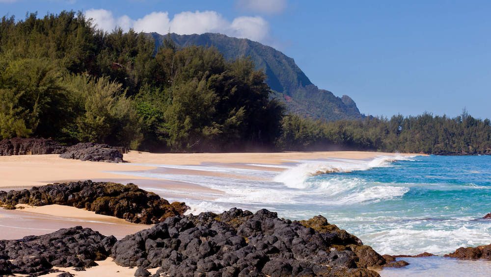 One of the top things to do in Hanalei Bay is head over to Lumahai Beach, one of the most beautiful beaches on Kauai. Image of a beach with black rocks in the foreground and mountains in the background.