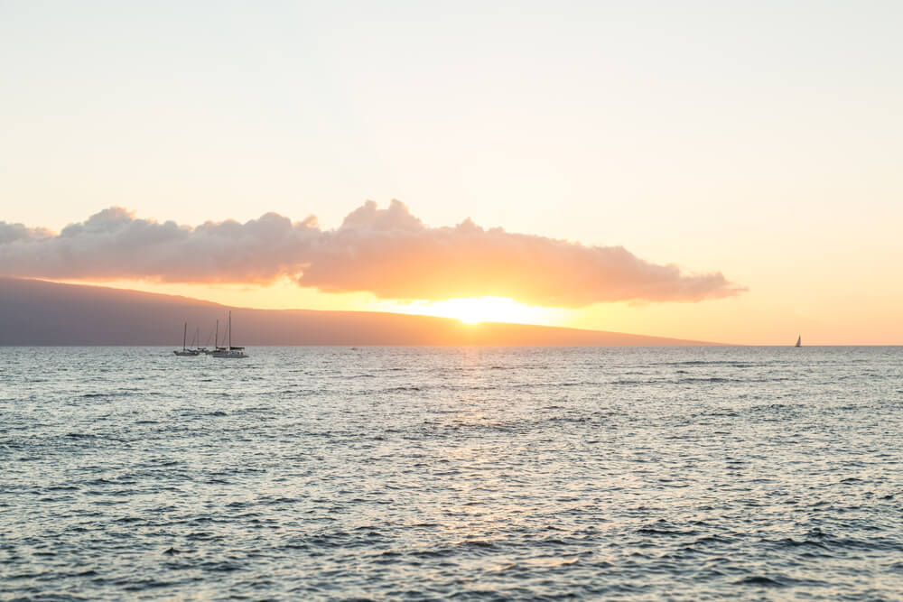 Hop on a Maui sunset cruise to see beautiful views of the sun setting behind the ocean. Image of a Lahaina sunset across the ocean with a few boats in the water.