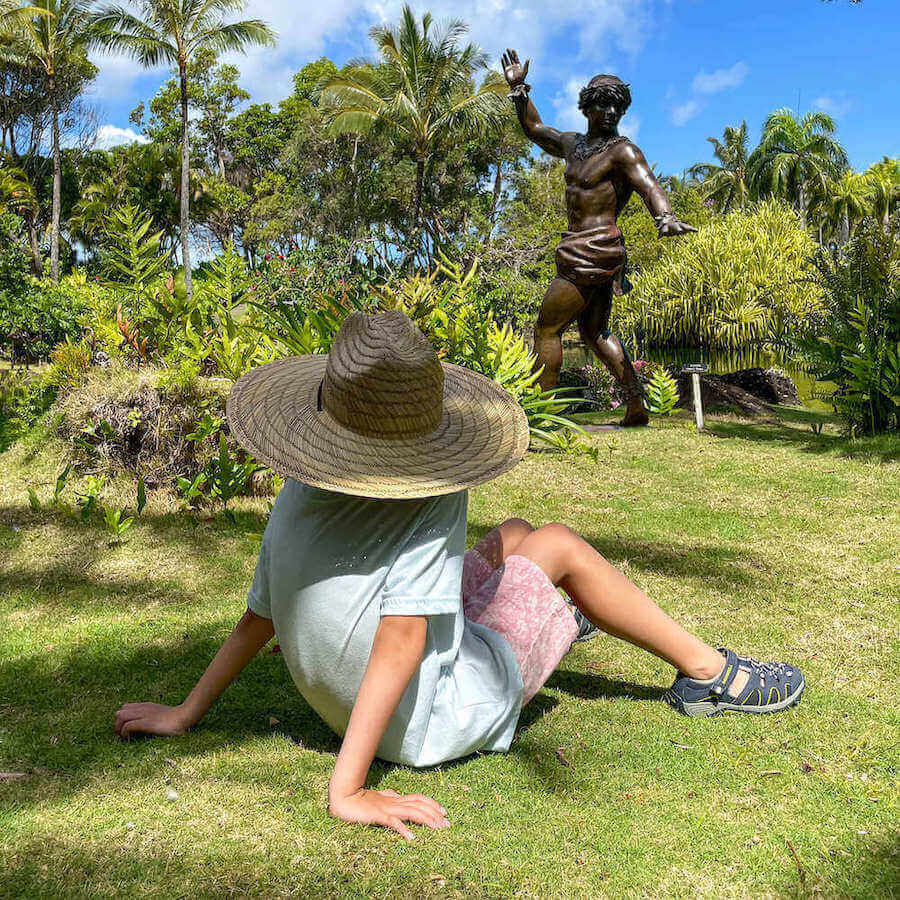 One of the best things to do near Hanalei is visit the Na Aina Kai Botanical Gardens in Kilauea. Image of a boy wearing a large straw hat sitting in the middle of a lush garden on Kauai.