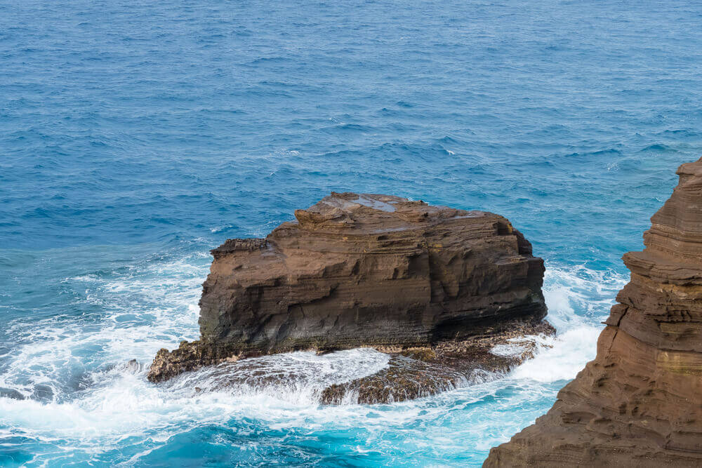 Looking for hidden gems in Oahu? Check out the Spitting Cave. Image of a huge brown lava rock in the blue ocean in Hawaii.