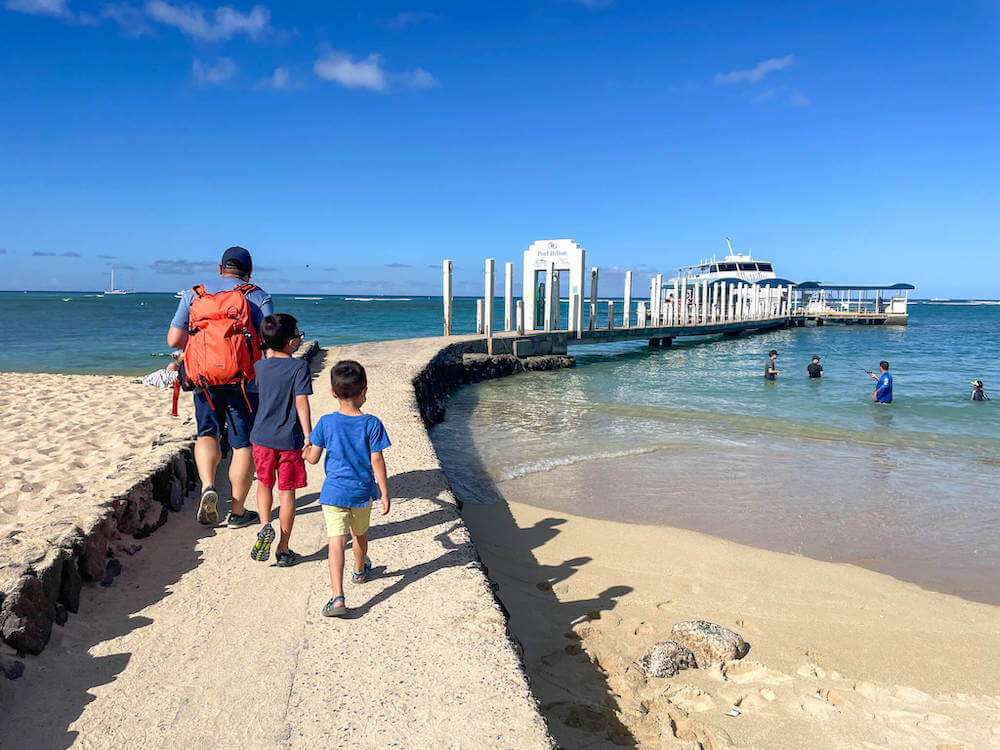 Find out how to get to the Waikiki Atlantis Submarine by top Hawaii blog Hawaii Travel with Kids. Image of a dad and two boys walking on a beach path to a boat.