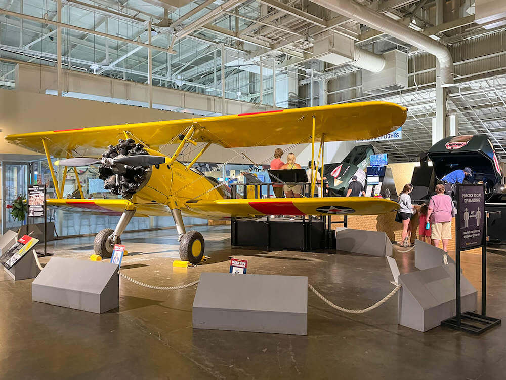 Image of a bright yellow WWII airplane, the Boeing N2S-3 Stearman with flight simulators in the background. These are located in Hangar 37 at the Pearl Harbor Aviation Museum.