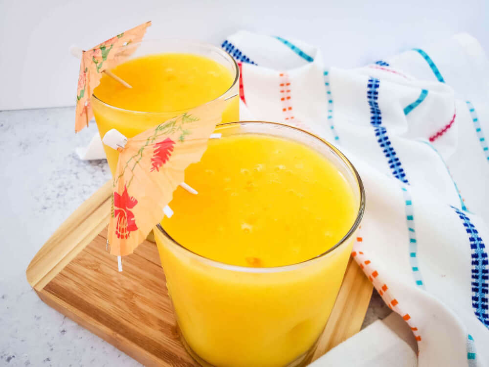 Find out how to make this easy frozen mango margarita recipe by top Hawaii blog Hawaii Travel with Kids. Image of two tumblers filled with an orange colored cocktail topped with little umbrellas.