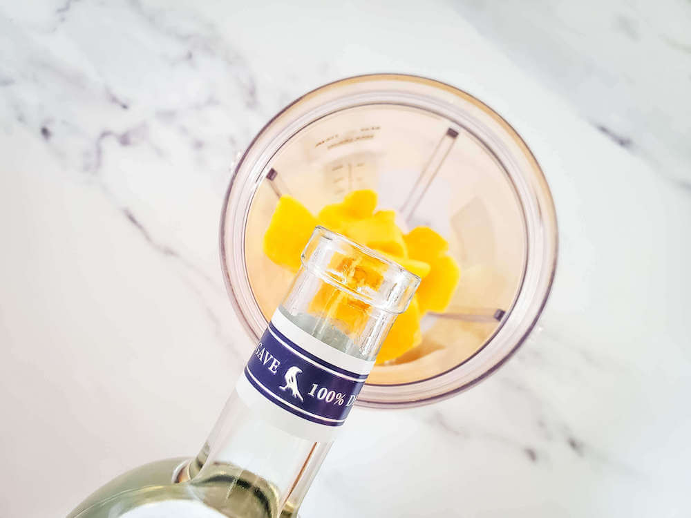 Add in your mango margarita ingredients and blend. Image of someone pouring alcohol into the blender over frozen mango chunks.