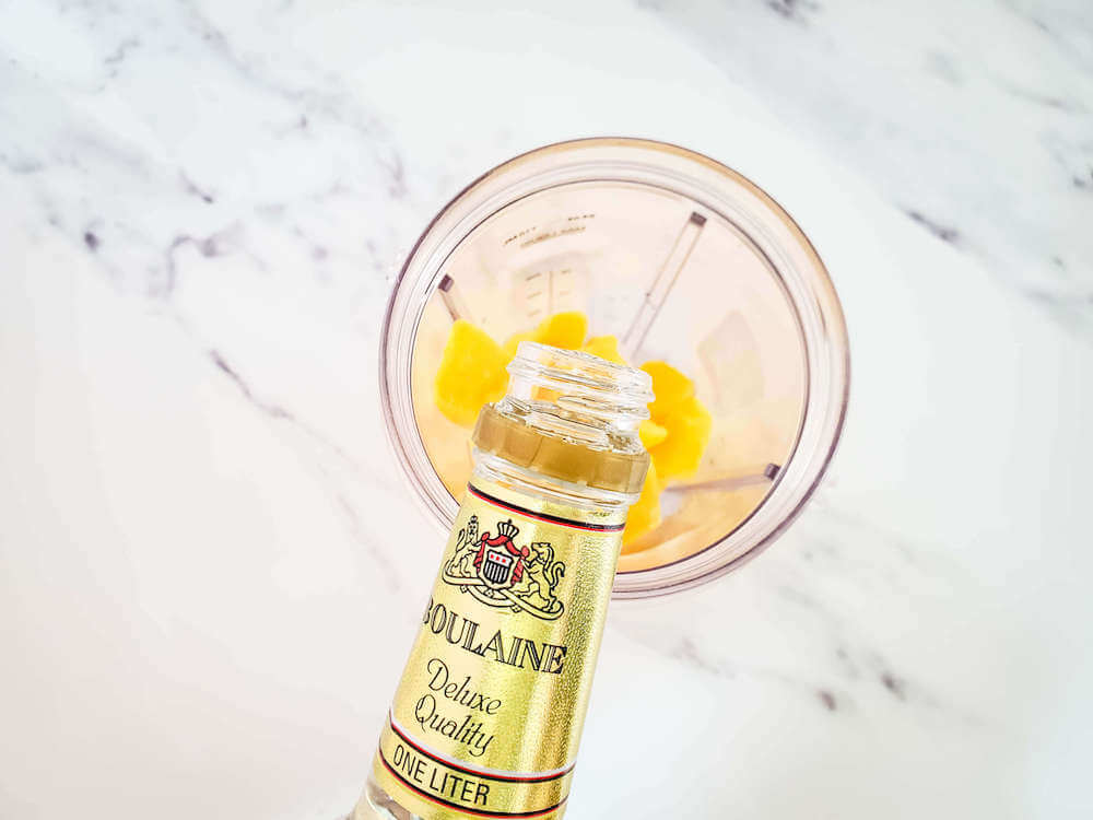 Add all your frozen mango margarita ingredients into the blender. Image of someone pouring alcohol over frozen mango and ice in a blender with a white background.