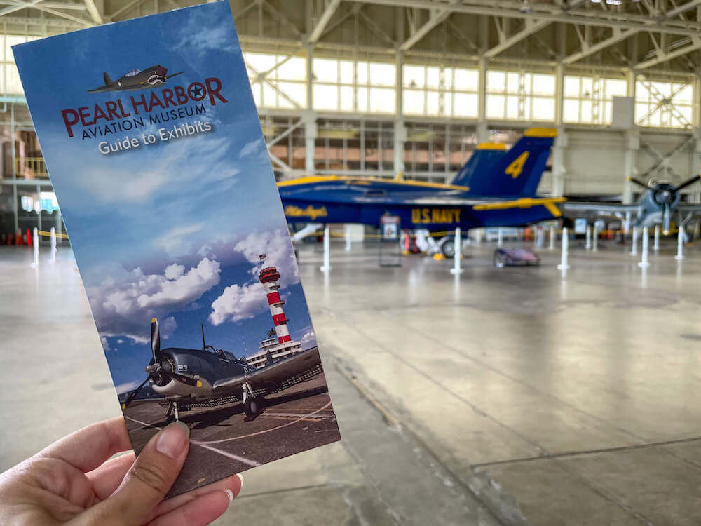 Image of a woman holding a Pearl Harbor Aviation Museum brochure with a Blue Angel airplane in the background.