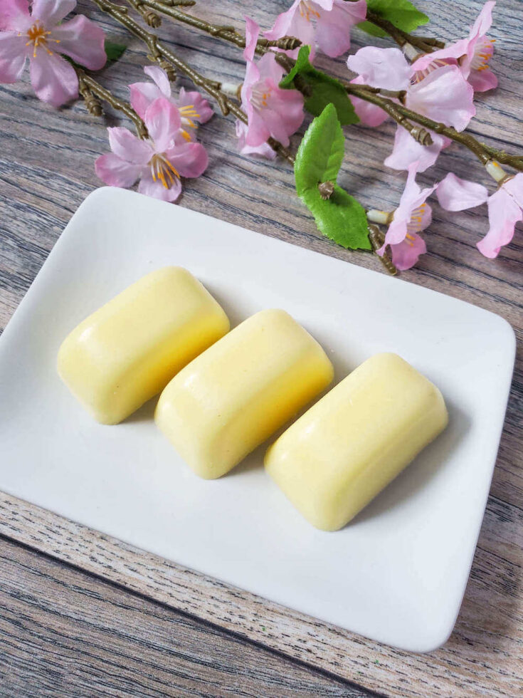 Find out how to make these pineapple sugar scrub bars from top Hawaii blog Hawaii Travel with Kids. Image of three yellow sugar scrub bars on a white plate with stems of pink flowers.