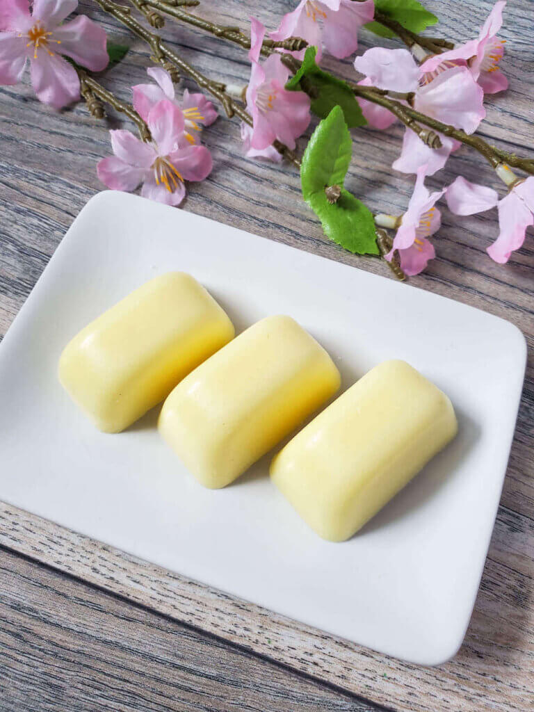 Find out how to make these pineapple sugar scrub bars from top Hawaii blog Hawaii Travel with Kids. Image of three yellow sugar scrub bars on a white plate with stems of pink flowers.