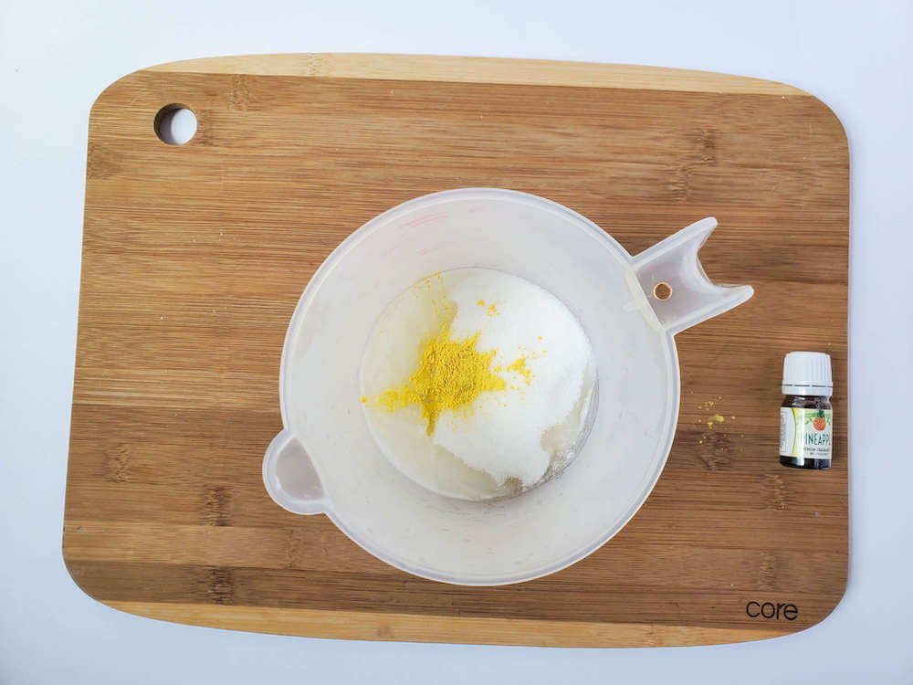 Add in the sugar and mica powder to make sugar scrub bars. Image of a white liquid measuring cup with soap, sugar, almond oil, and mica powder inside.