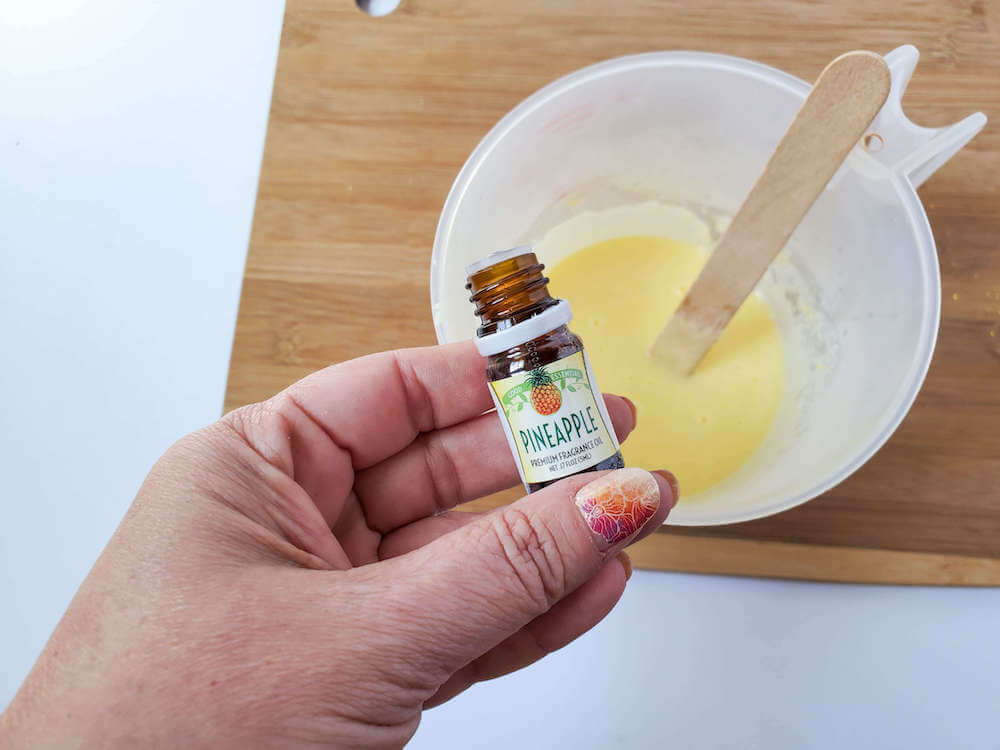 Add in the pineapple fragrance oil so your sugar scrub bars will smell tropical. Image of someone holding a little bottle of pineapple fragrance oil in front of a measuring cup.