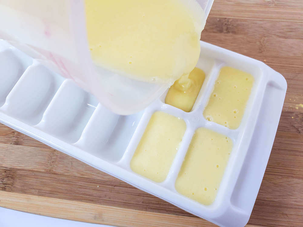 Pour your pineapple sugar scrub mixture into an ice cube tray. Image of someone pouring yellow liquid into a white ice cube tray.