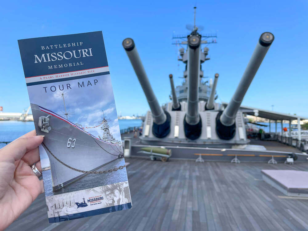 Remember to grab a tour map of the Battleship Missouri Memorial at Pearl Harbor. Image of a woman holding a tour map brochure with the USS Battleship Missouri in the background.