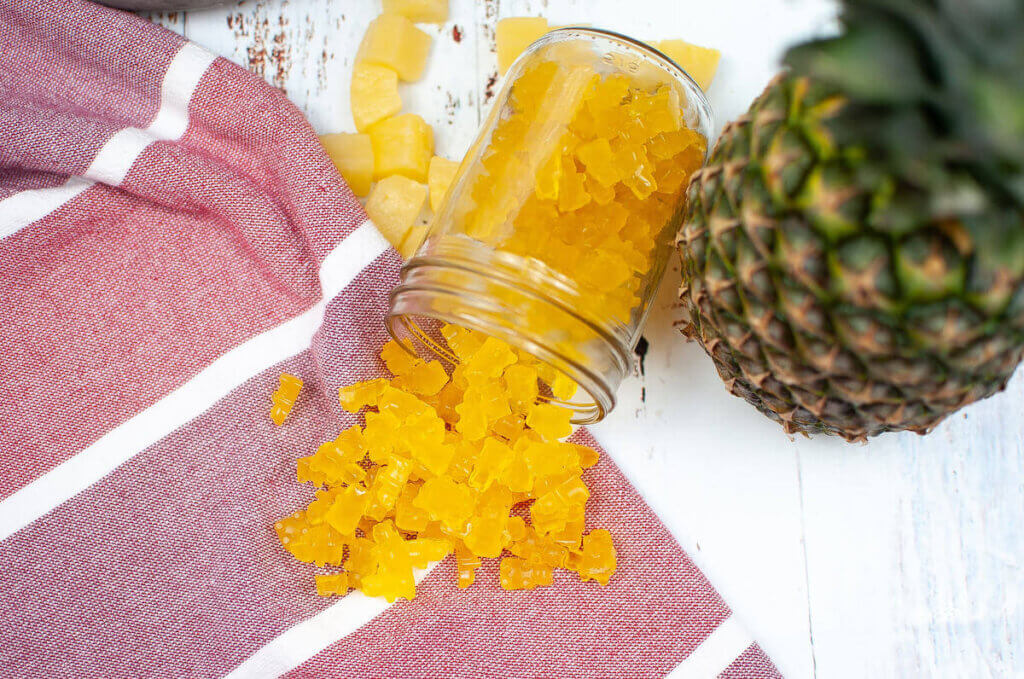 Image of a jar of pineapple gummy bears spilling onto a pink kitchen towel with a whole pineapple next to it.