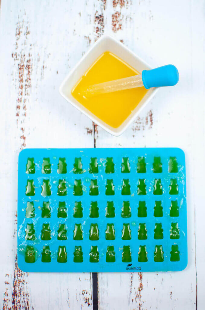 Fill a gummy bear mold with your pineapple gummy bear mixture by using a dropper. Image of a blue gummy bear mold, a dropper, and a bowl of yellow mixture.