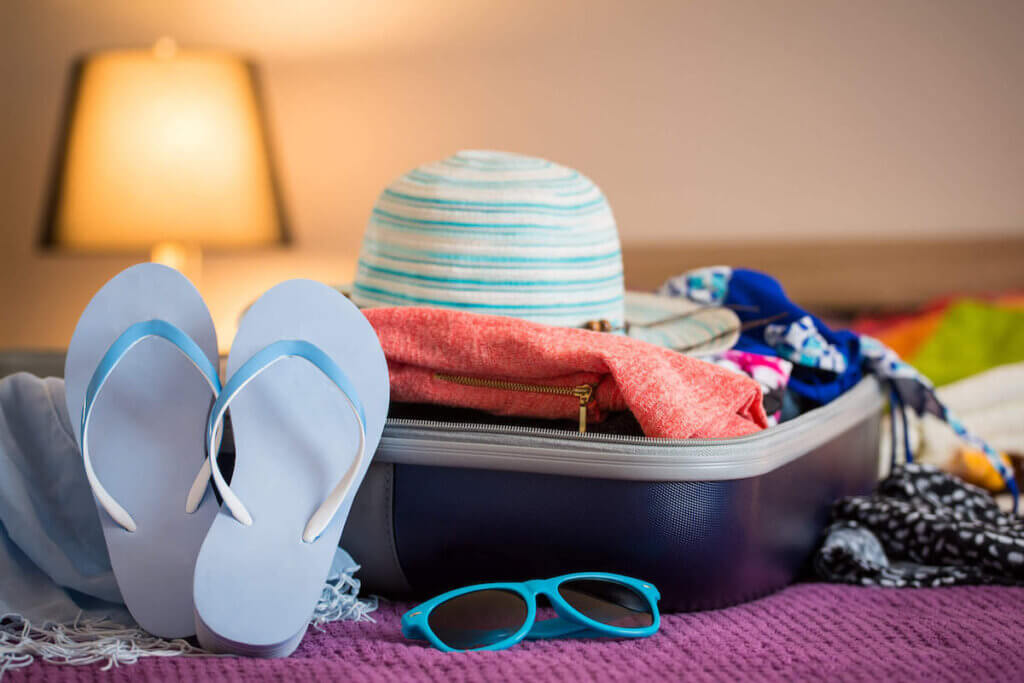 Your Hawaii packing list for October will look like any other month, but you might want to add a pair of pants and a light sweater. Image of a suitcase on a bed with blue flip flops, sunglasses, and a hat.