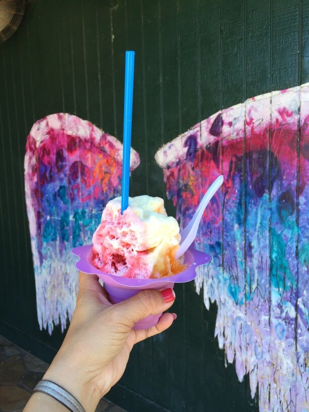 One of the best places to take photos on Oahu is the angel wings mural in Haleiwa. Image of a woman holding shave ice in front of angel wings.