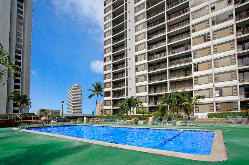One of the best places to stay on Oahu on a budget is the Aston at the Waikiki Banyan. Image of a pool on a green deck with hotel towers around it.