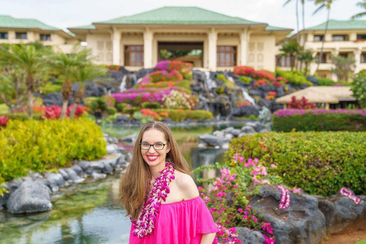 Read the full Grand Hyatt Kauai review by top Hawaii blog Hawaii Travel with Kids. Image of a woman wearing a pink dress standing in front of a waterfall and hotel