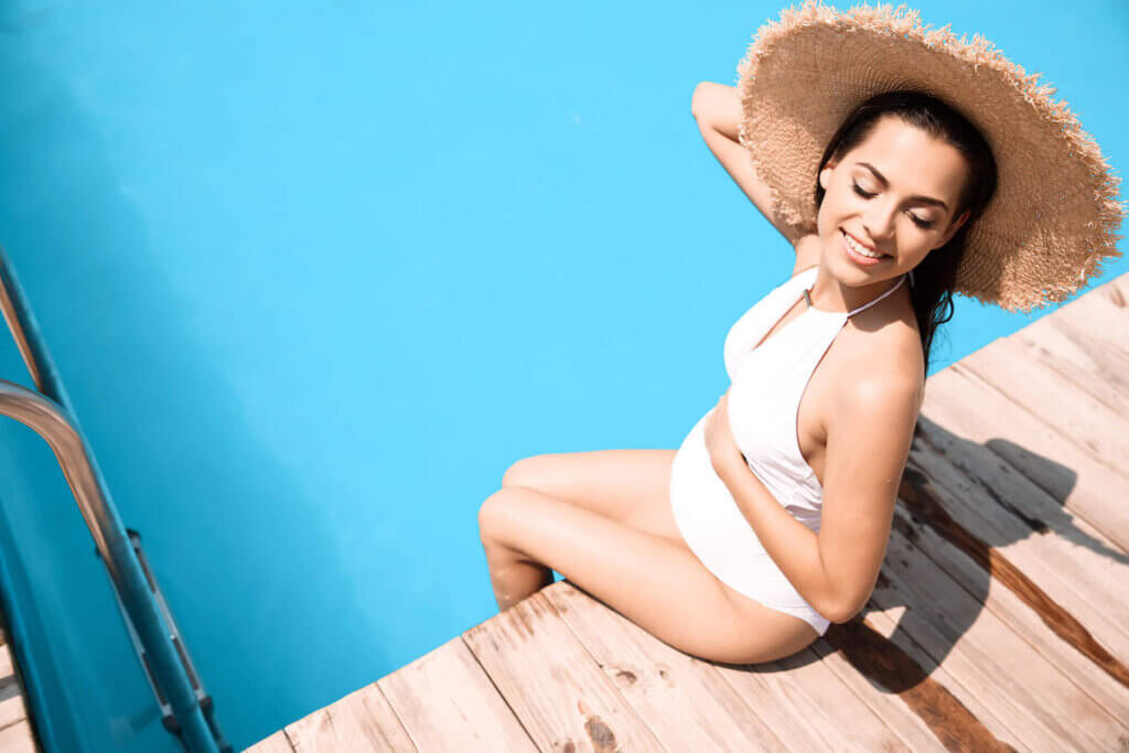 This Hawaii babymoon packing list includes everything to wear in Hawaii while pregnant. Image of of a pregnant woman wearing a white swimsuit and sun hat.