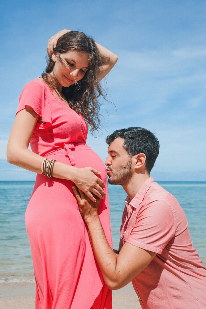 Image of a man kissing the pregnant belly of a woman wearing a pink maternity dress.