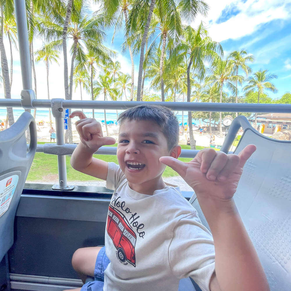 Image of a boy waving two shakas while sitting on top a double decker bus in Waikiki.