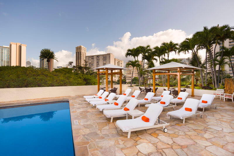 One of the cheapest places to stay on Oahu is the Luana Waikiki Hotel & Suites. Image of white lounge chairs with orange pillows next to a swimming pool in Hawaii.