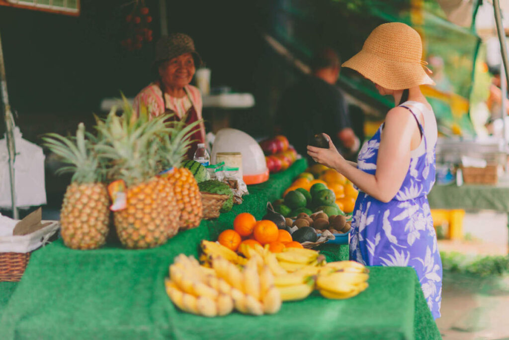 Find out the best Oahu farmers markets recommended by top Hawaii blog Hawaii Travel with Kids. Image of a woman buying tropical fruit at a Hawaii farmers market.