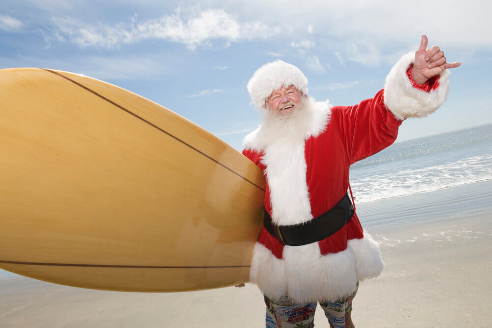 One of the best Christmas traditions in Hawaii is that you can spot surfing Santa! Image of Santa holding a surfboard and waving a shaka sign in Hawaii.