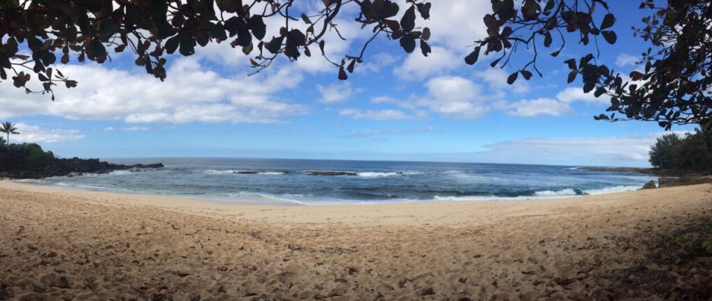 Three Tables Beach is one of the best North Shore Hawaii beaches. Image of a panoramic view of a sandy beach and ocean with trees on the sides.