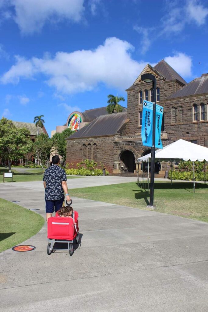 It's helpful to bring a wagon when visiting Bishop Museum with kids. Image of a dad pulling a red wagon with two toddlers outside of Bishop Museum in Hawaii.