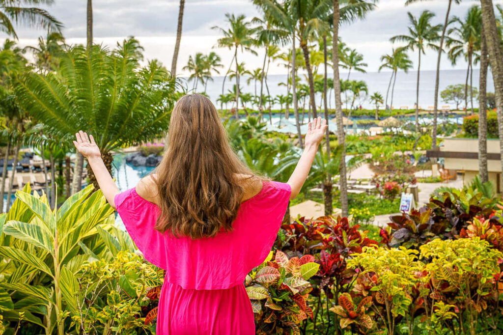 Read my full Grand Hyatt Kauai review by top Hawaii blog Hawaii Travel with Kids. Image of a woman with outstretch arms facing the pool area at the Grand Hyatt Kauai.