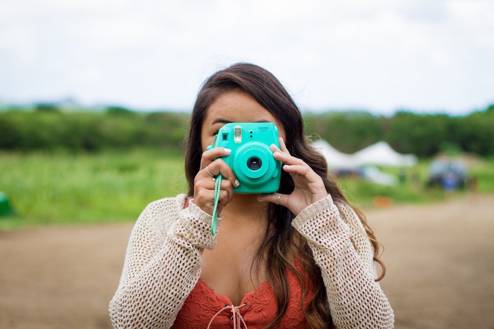 Find out the best travel quotes about Hawaii for Instagram by top Hawaii blog Hawaii Travel with Kids. Image of a woman taking a photo with a blue Polaroid camera in Hawaii.