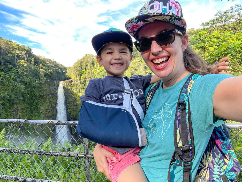 Image of a woman wearing a hat that says Aloha holding a boy with a sling in front of Akaka Falls on the Big Island of Hawaii.