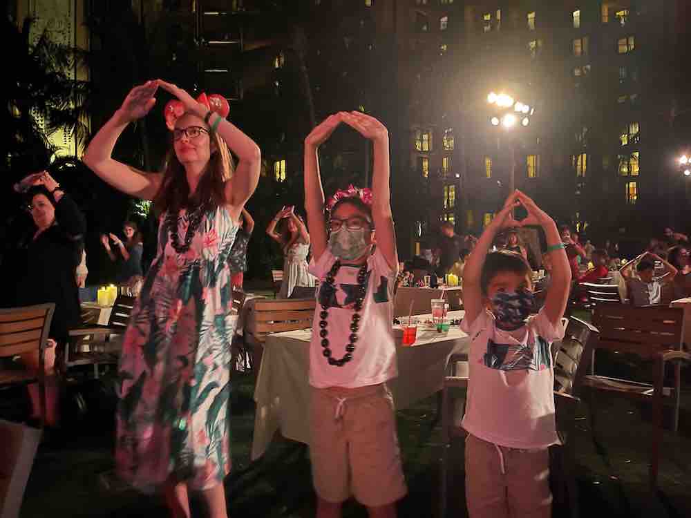 Image of a mom and two boys dancing hula at night next to luau tables.